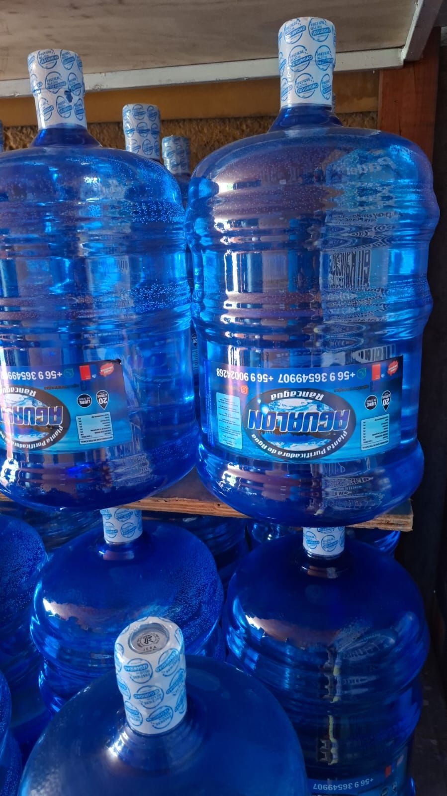 Grandes bidones de agua azules apilados en un área de almacenamiento, algunos con tapones blancos.
