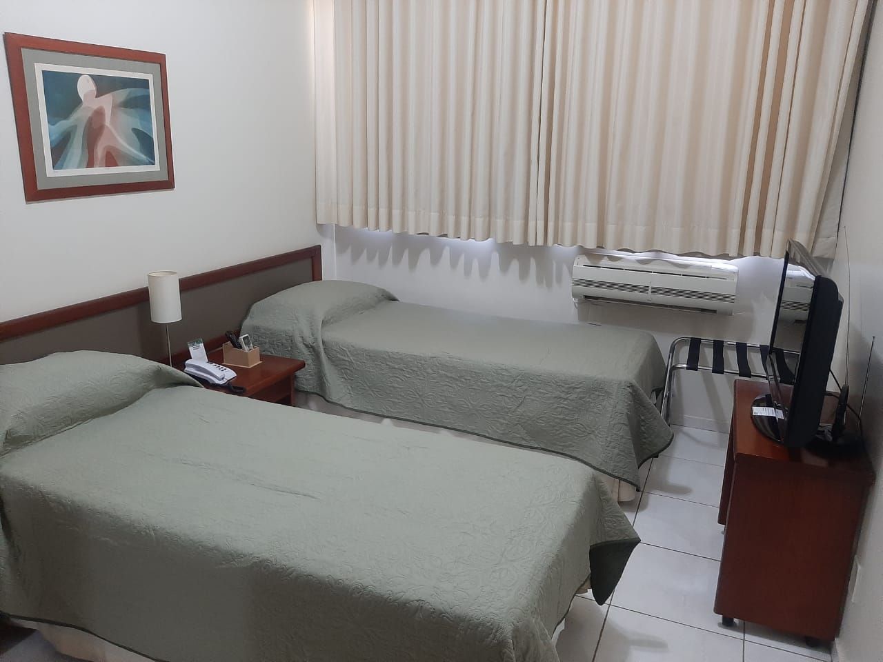 Um quarto de hotel com duas camas de solteiro com roupa de cama verde-sálvia, uma obra de arte emoldurada na parede e uma televisão sobre uma cômoda de madeira.