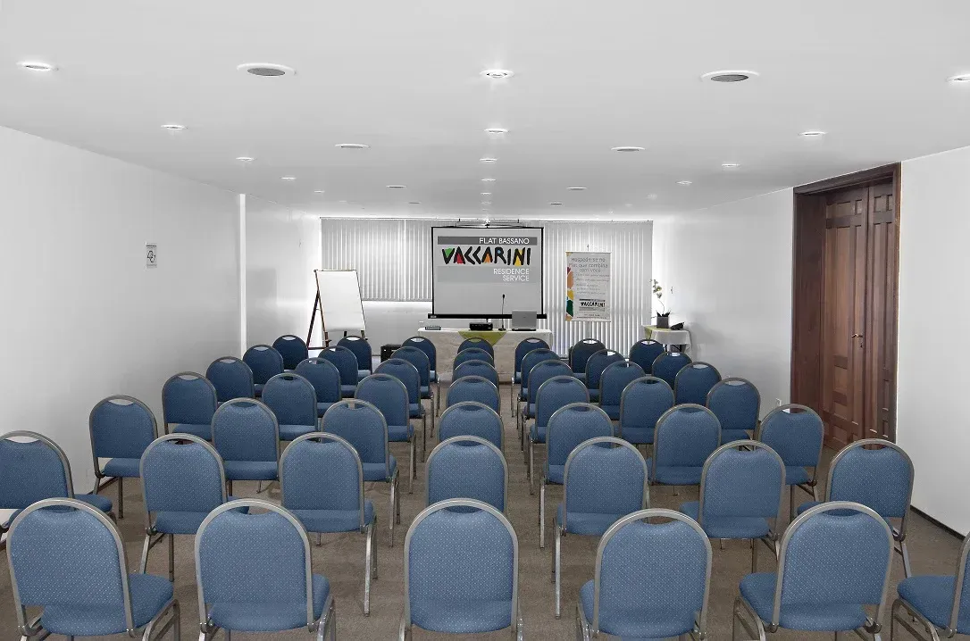 Uma sala de conferências vazia com fileiras de cadeiras azuis voltadas para uma tela de projeção e uma parede branca com uma porta de madeira.
