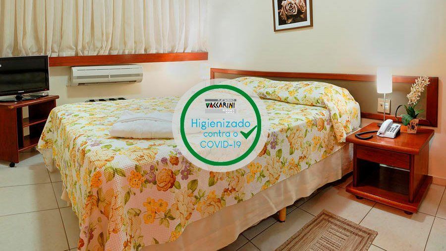 Um quarto de hotel com uma cama com roupa de cama floral, uma mesa de cabeceira de madeira com um abajur e um telefone, e uma TV sobre um suporte.