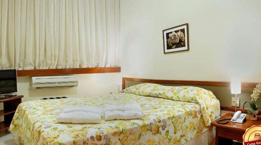 Um quarto de hotel com uma cama queen-size com uma colcha floral amarela, cortinas brancas, uma mesa de cabeceira e um quadro na parede.