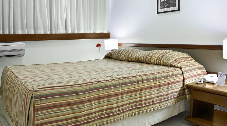 Um quarto com uma cama com colcha listrada em tons de bege e marrom, cortinas brancas e uma mesa de cabeceira de madeira.
