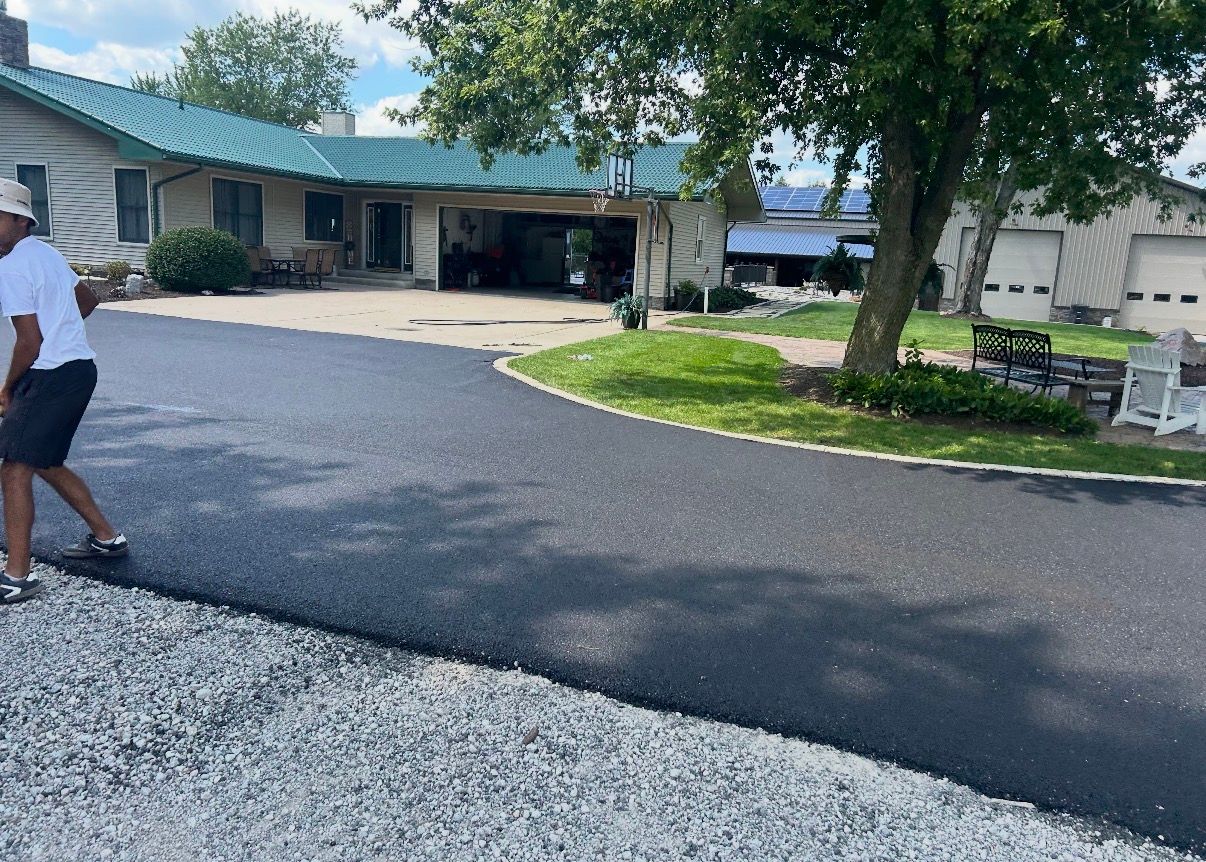 Asphalt driveway between brick building and gray-sided house. Wet surface, sidewalk in foreground.