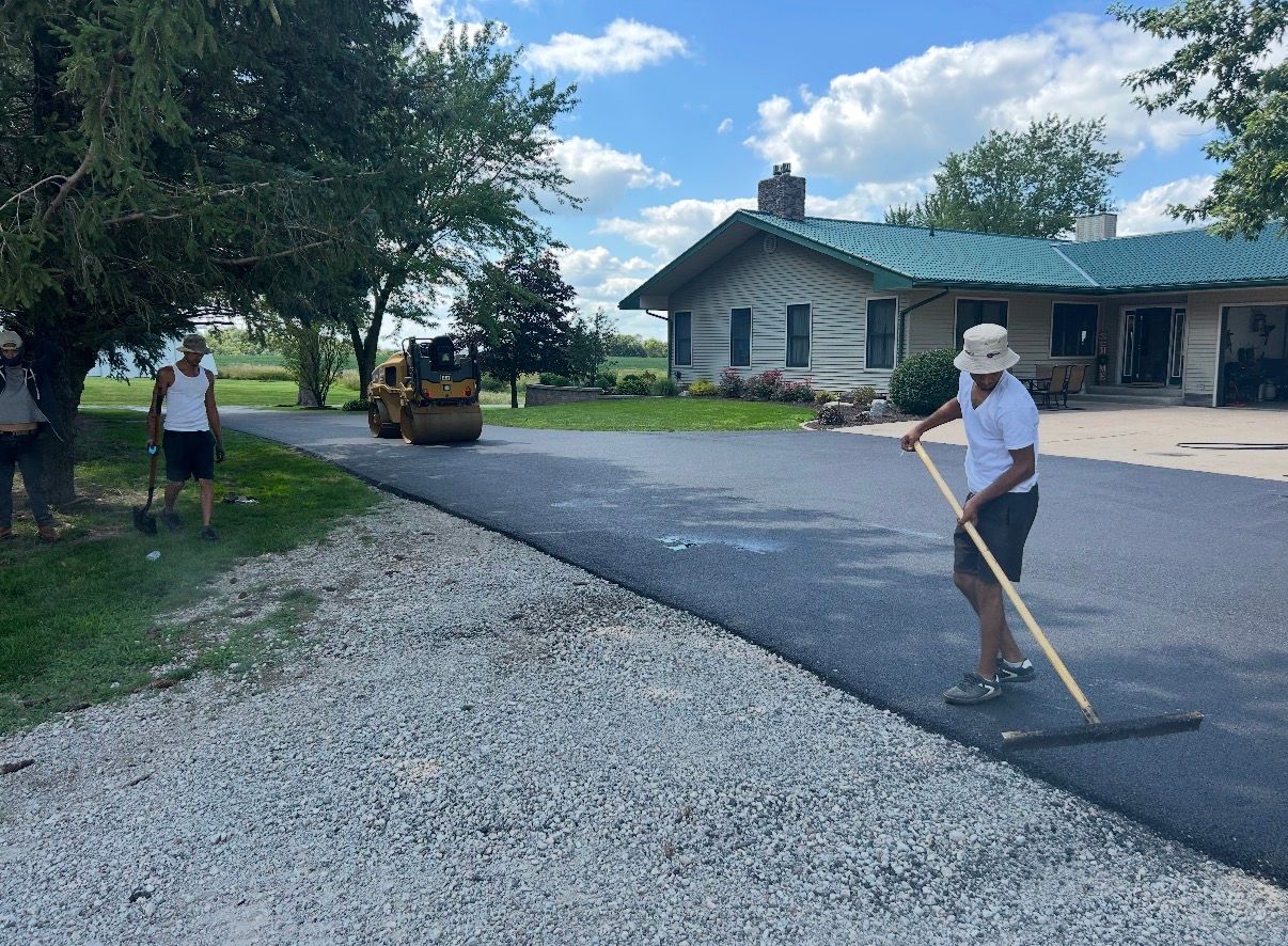 Asphalt driveway between brick building and gray-sided house. Wet surface, sidewalk in foreground.