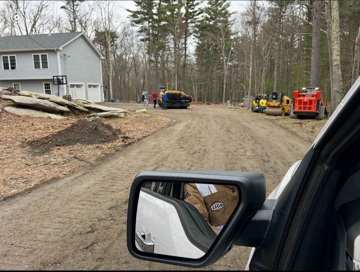 Asphalt driveway between brick building and gray-sided house. Wet surface, sidewalk in foreground.
