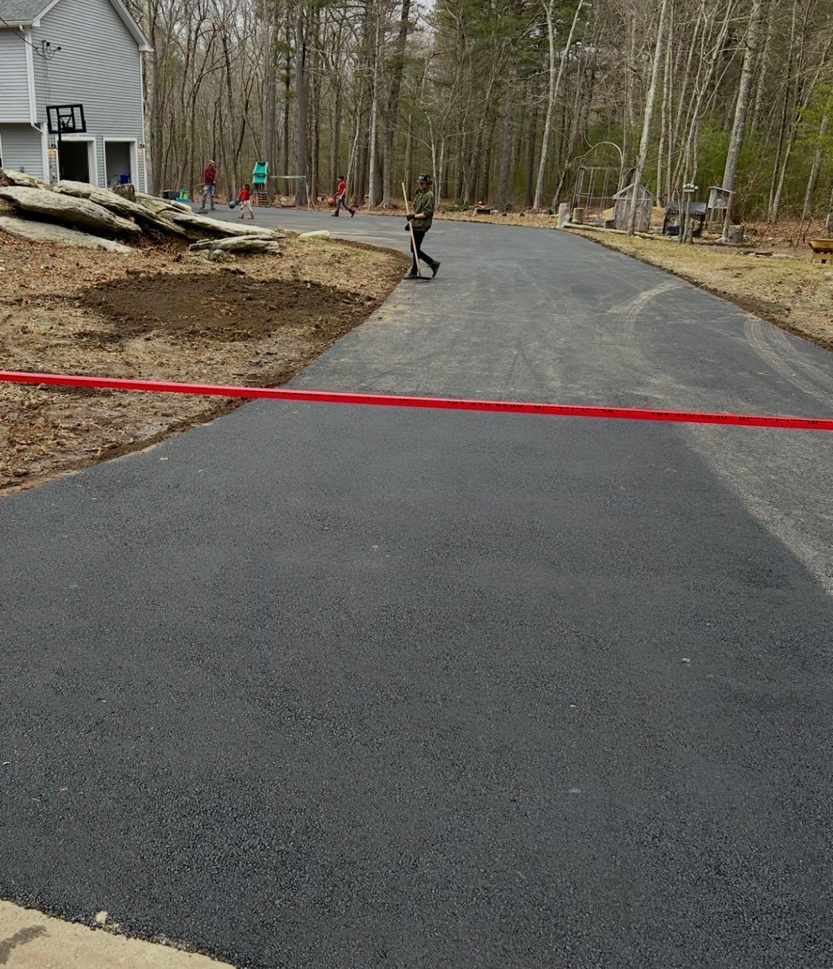 Asphalt driveway between brick building and gray-sided house. Wet surface, sidewalk in foreground.