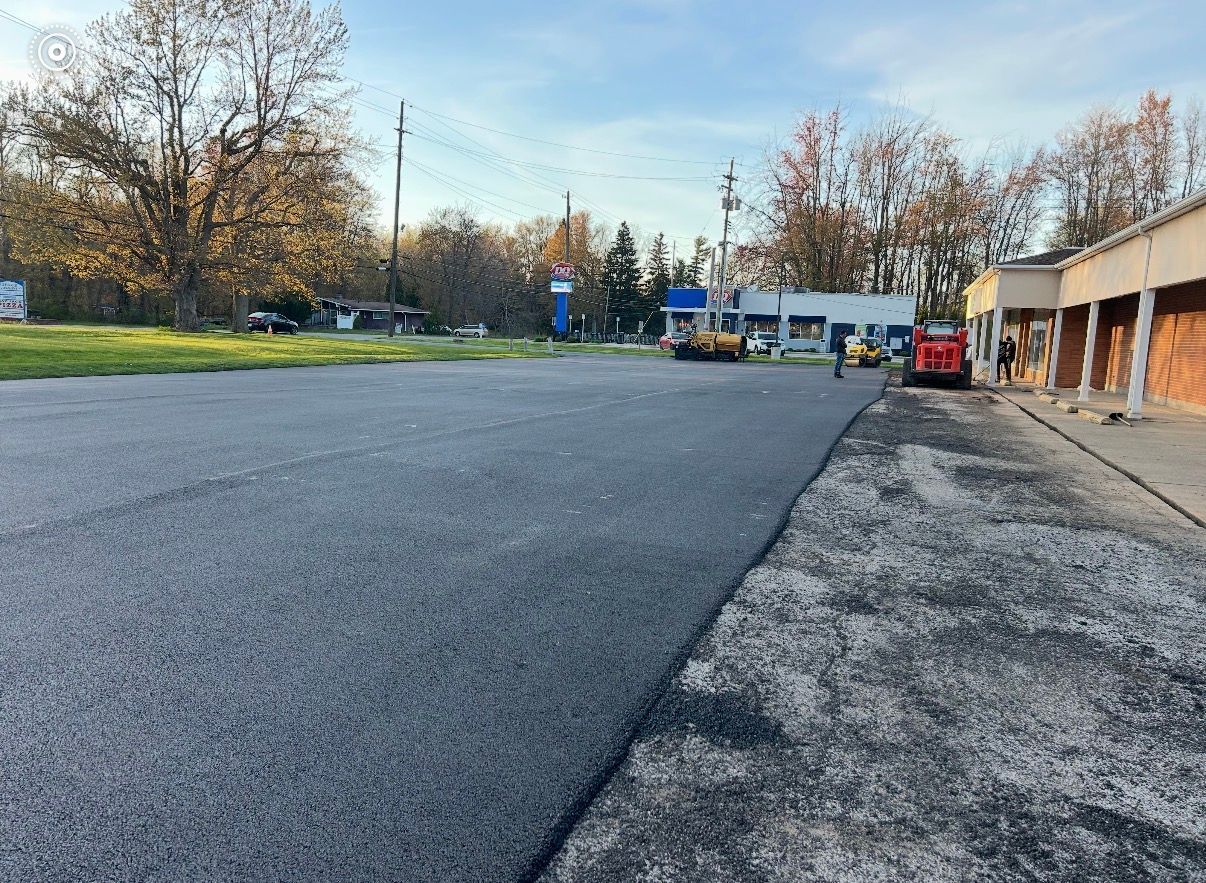Newly paved black asphalt driveway in front of a blue-gray house with a white garage door, yellow markings, and a white sign.