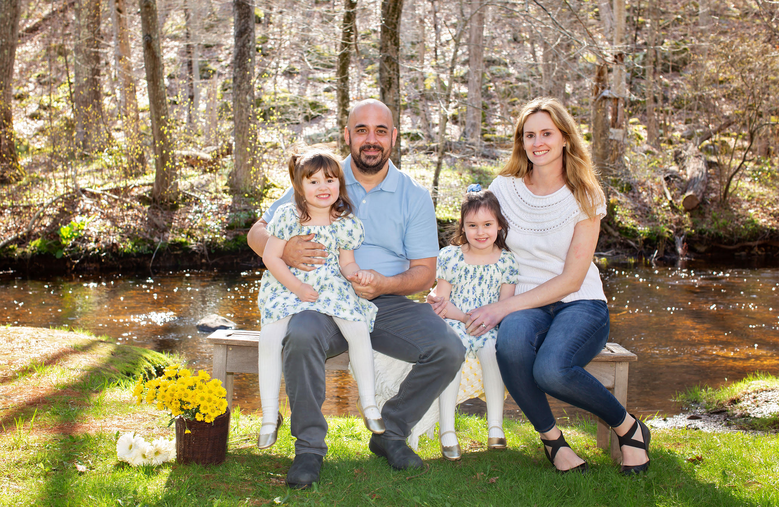 Family seated on chairs by a creek in a sunny wooded park, smiling at the camera.