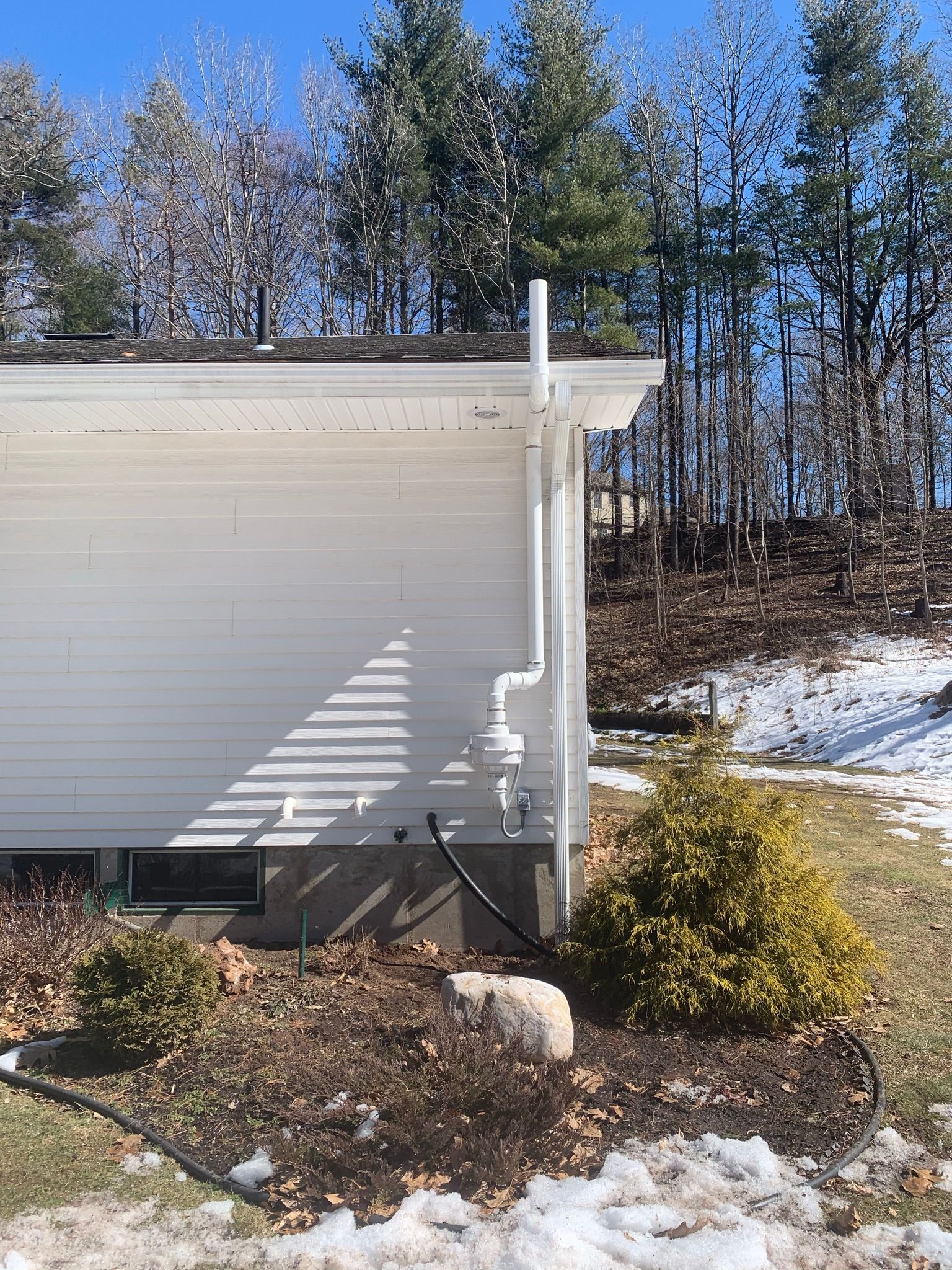 Side of a white house with downspout, snowy yard, and trees in the background.