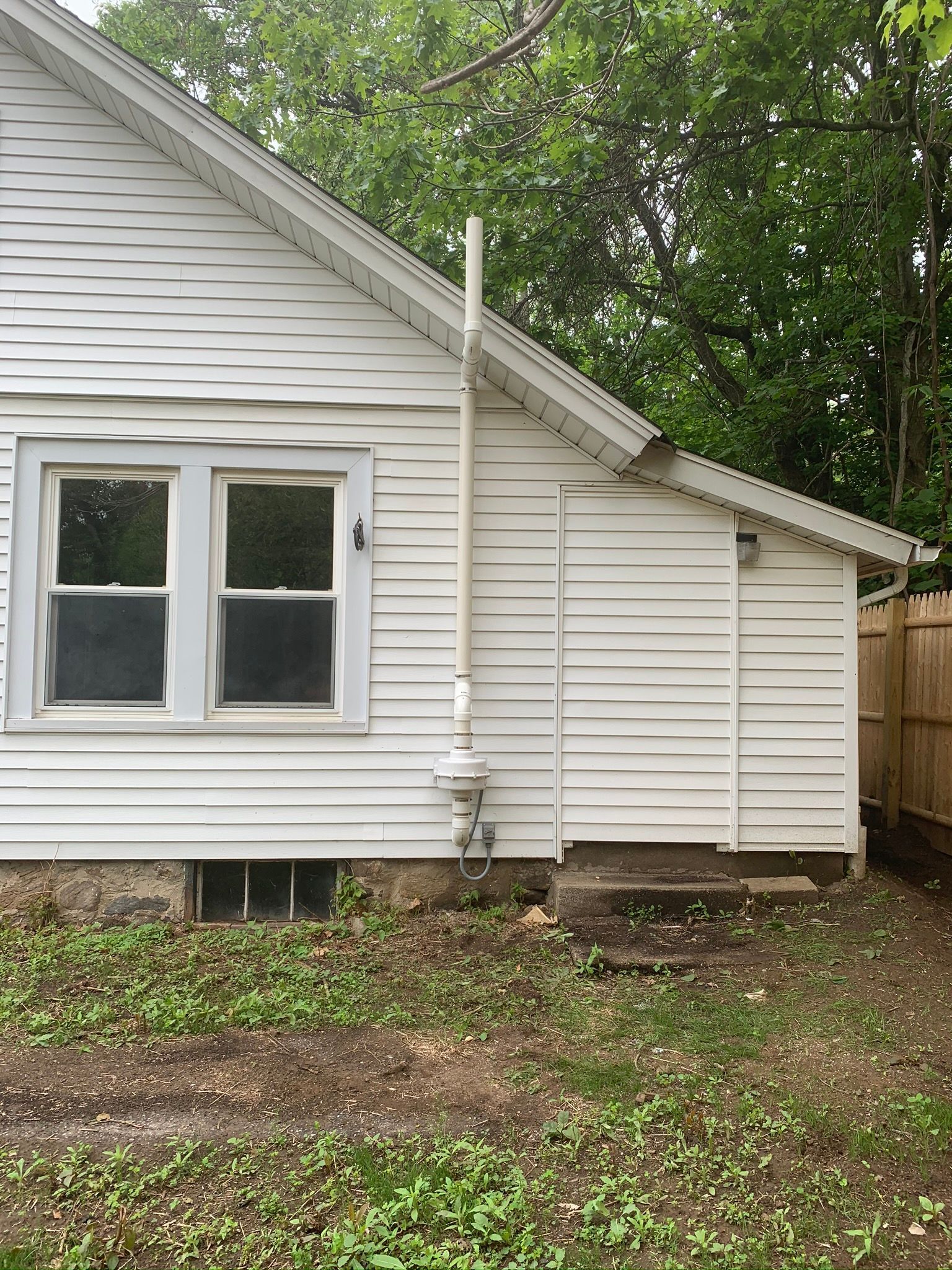 Side of a white house with a double window, downspout, and trees in the yard.