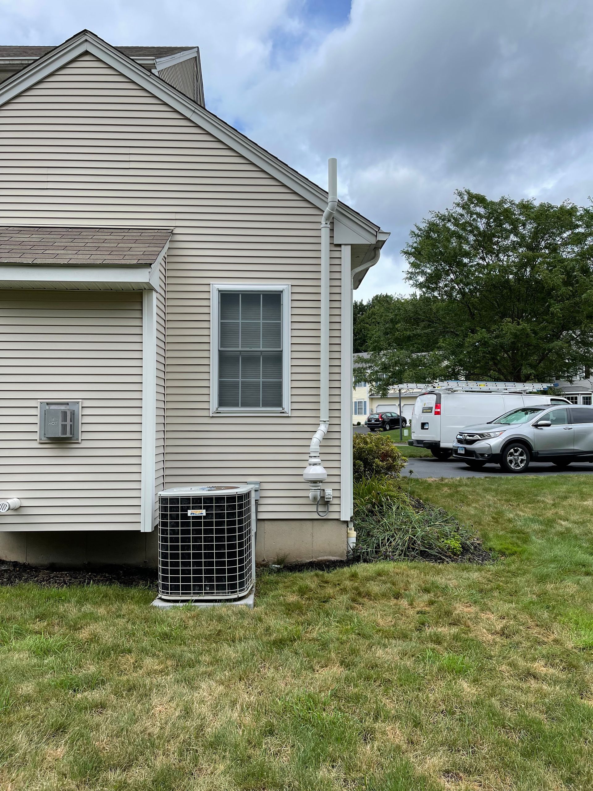 Side of beige house with window unit AC, grassy yard, and parked cars under cloudy sky