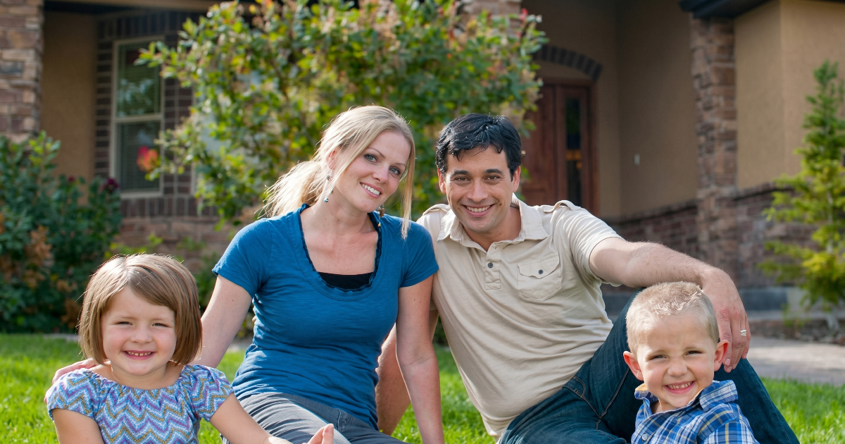 Family sitting on grass outside a house, smiling at the camera.