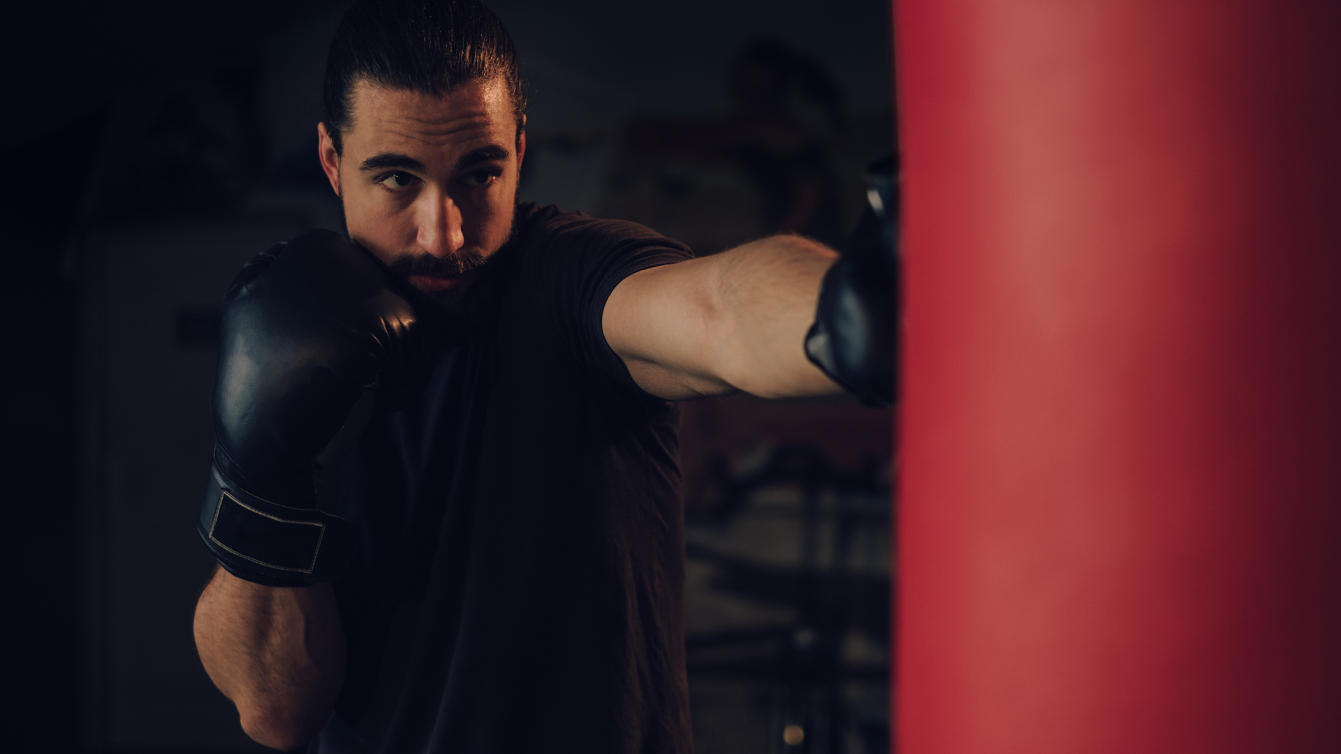 Boxer in black gloves punches a red heavy bag. Dark gym setting.