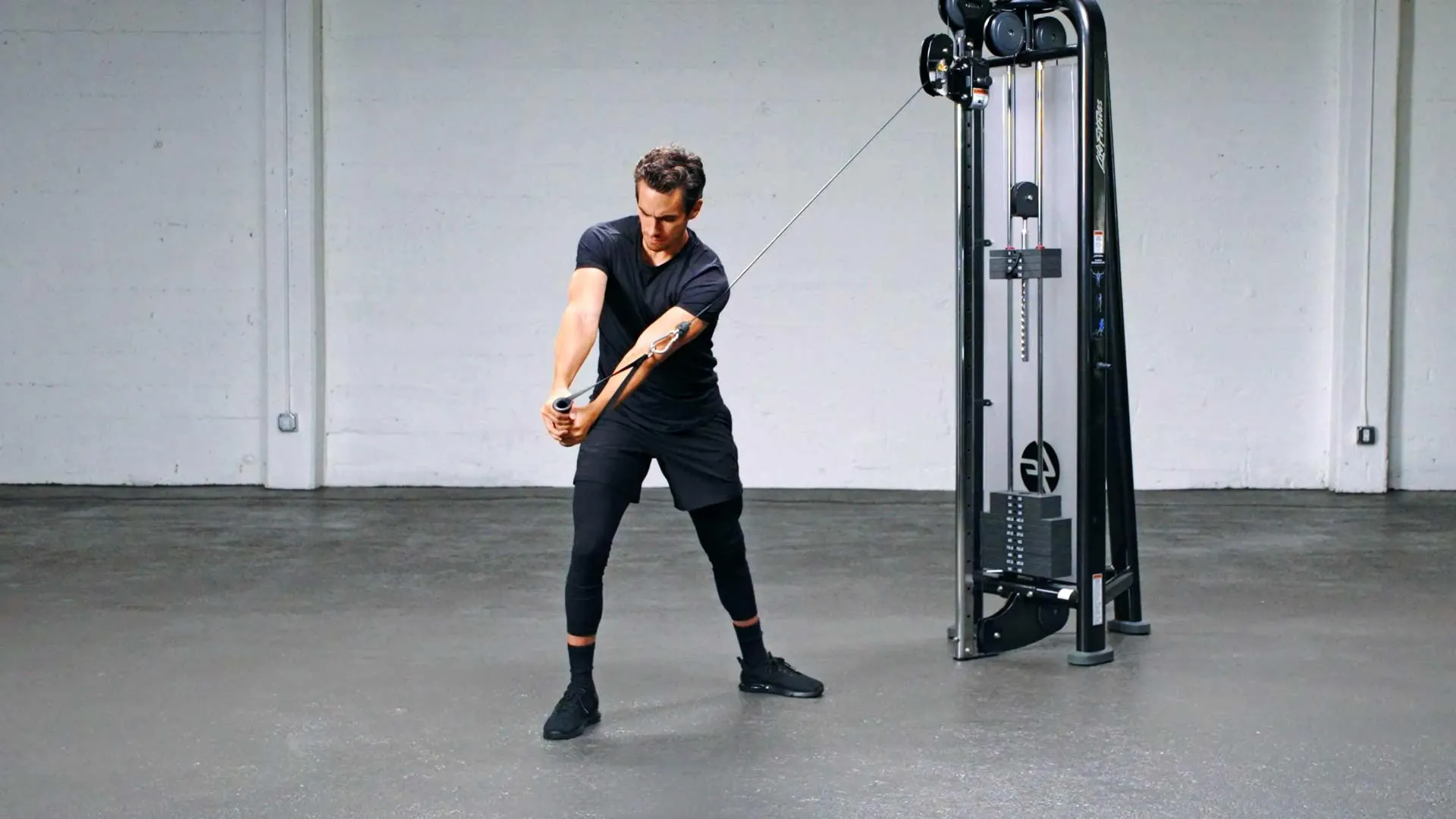 Person swinging a cable machine handle in a gym studio, standing on a gray floor near equipment.