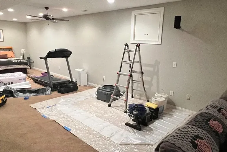 Basement room being renovated, with treadmill, ladder, paint supplies, and a couch covered in plastic.