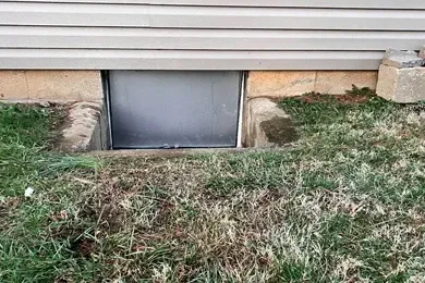 Basement window well with a gray cover under house siding, bordered by grass and dirt.