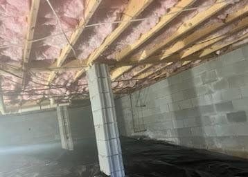 Unfinished basement with exposed pink insulation, wood joists, gray cinderblock walls, and a support column.