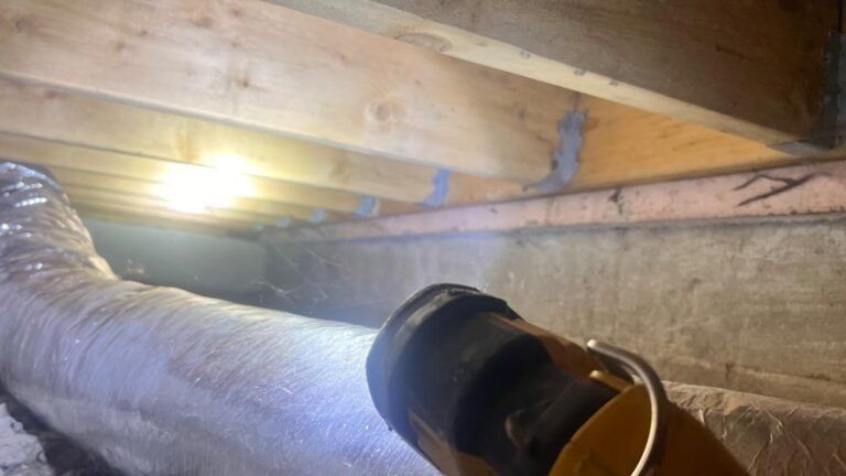 Inside a crawlspace, a flexible silver duct runs under wooden beams with a light glowing overhead.