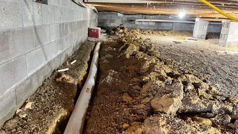 Crawl space under a house with exposed pipes, dirt floor, and muddy excavation along the foundation wall