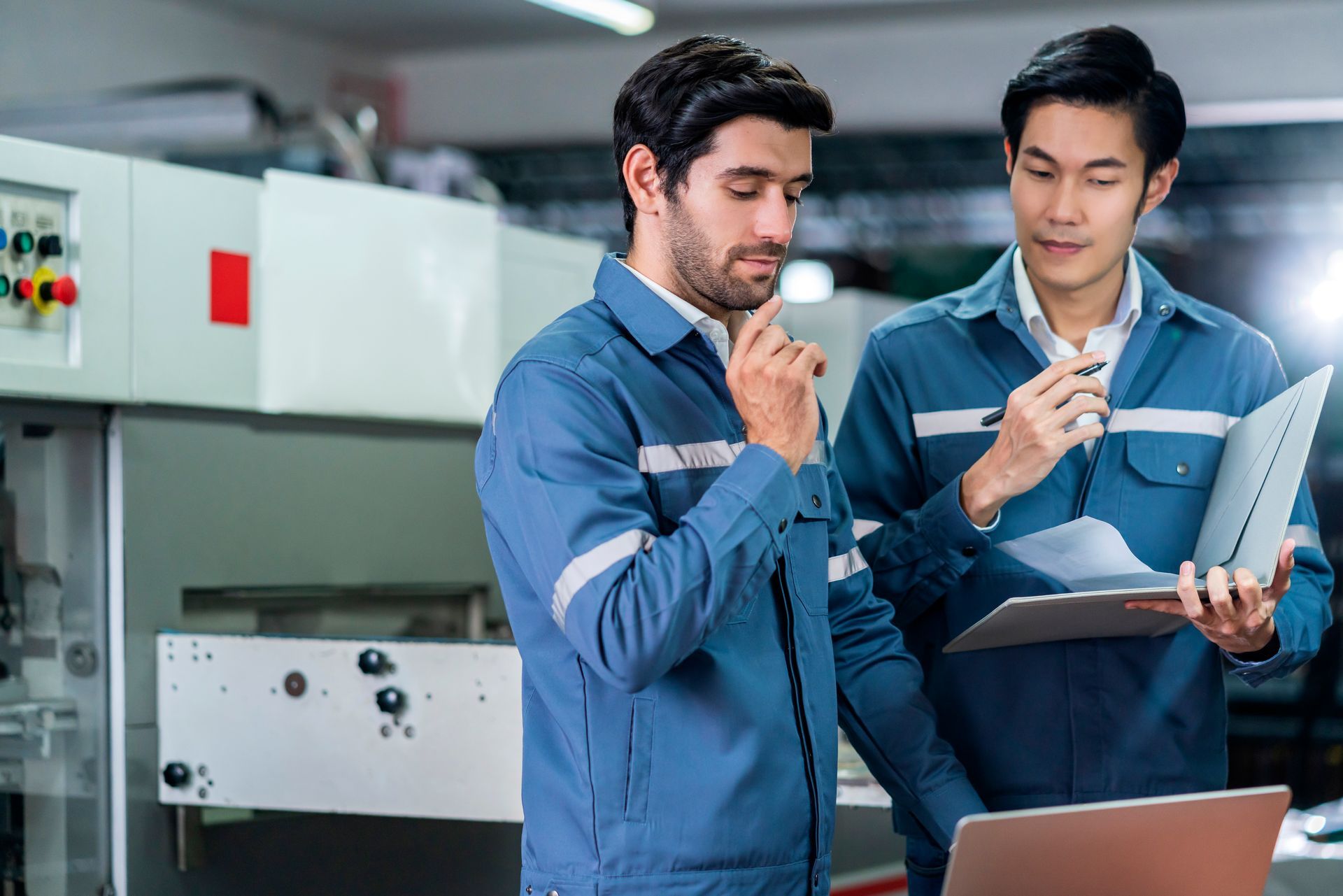 Two men in blue work uniforms examining a laptop and paperwork in a factory setting.