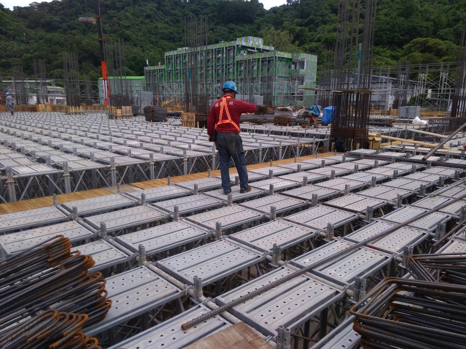 Construction worker on a scaffolding platform at a construction site, pointing; steel rebar and building under construction in the background.