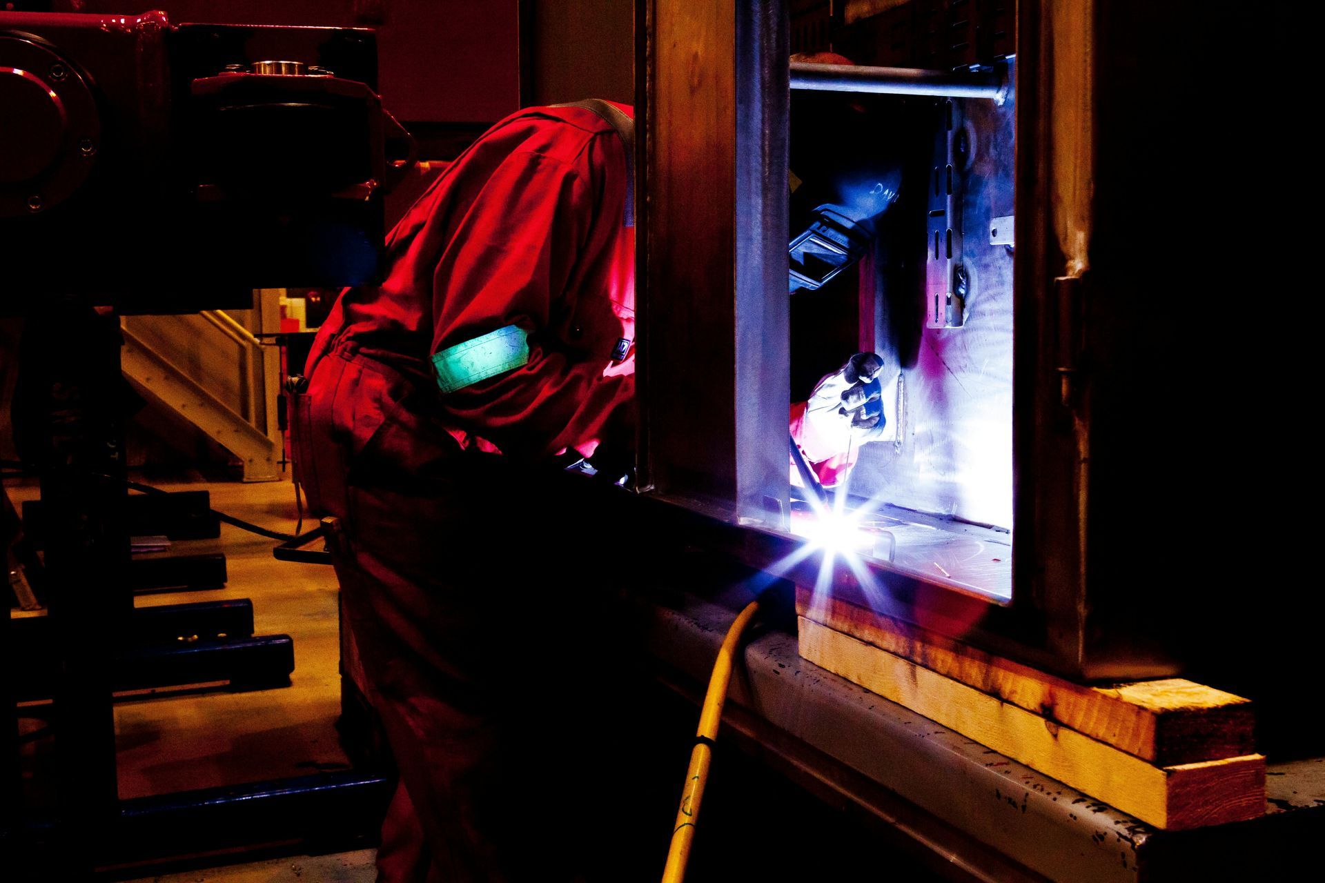 Welder in red protective suit working on metal, sparks flying in dimly lit space.