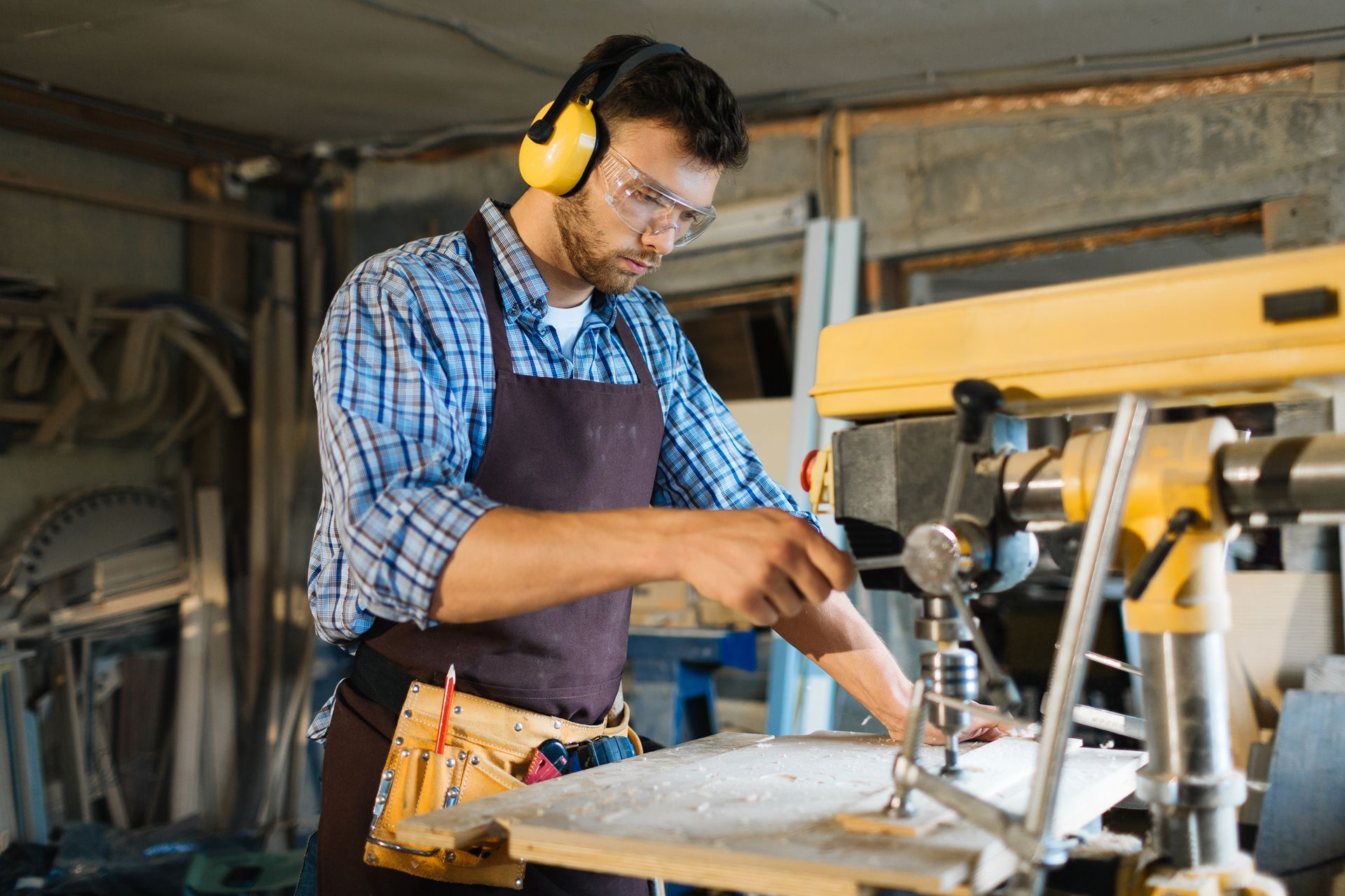 Carpenter in a workshop using a drill press, wearing safety glasses, ear protection, and an apron.