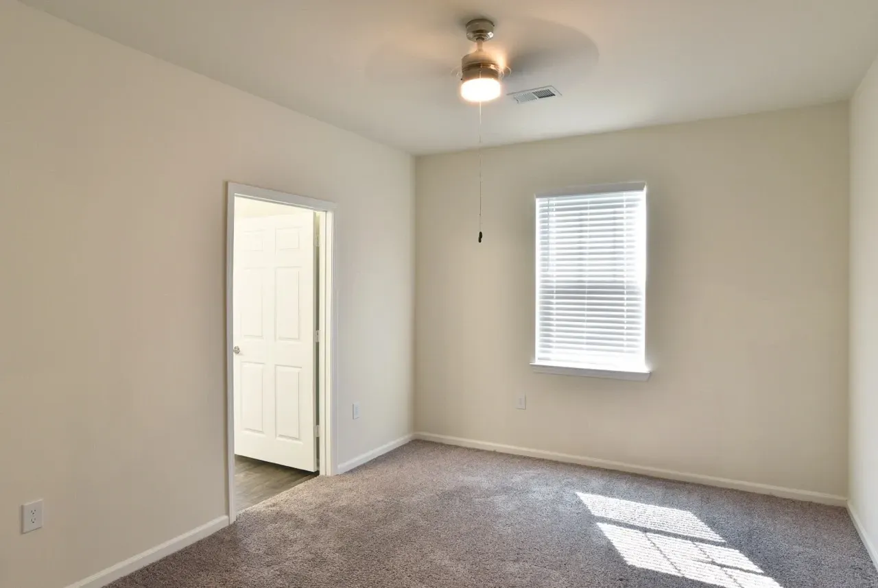 Empty bedroom with beige walls, carpet, ceiling fan, and a window with blinds.