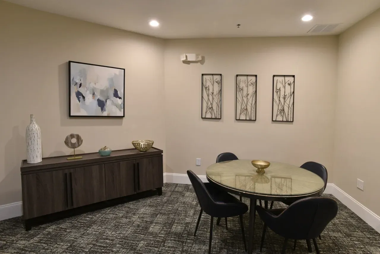 Well-lit communal lounge with a round glass table, four dark chairs, and wall art.