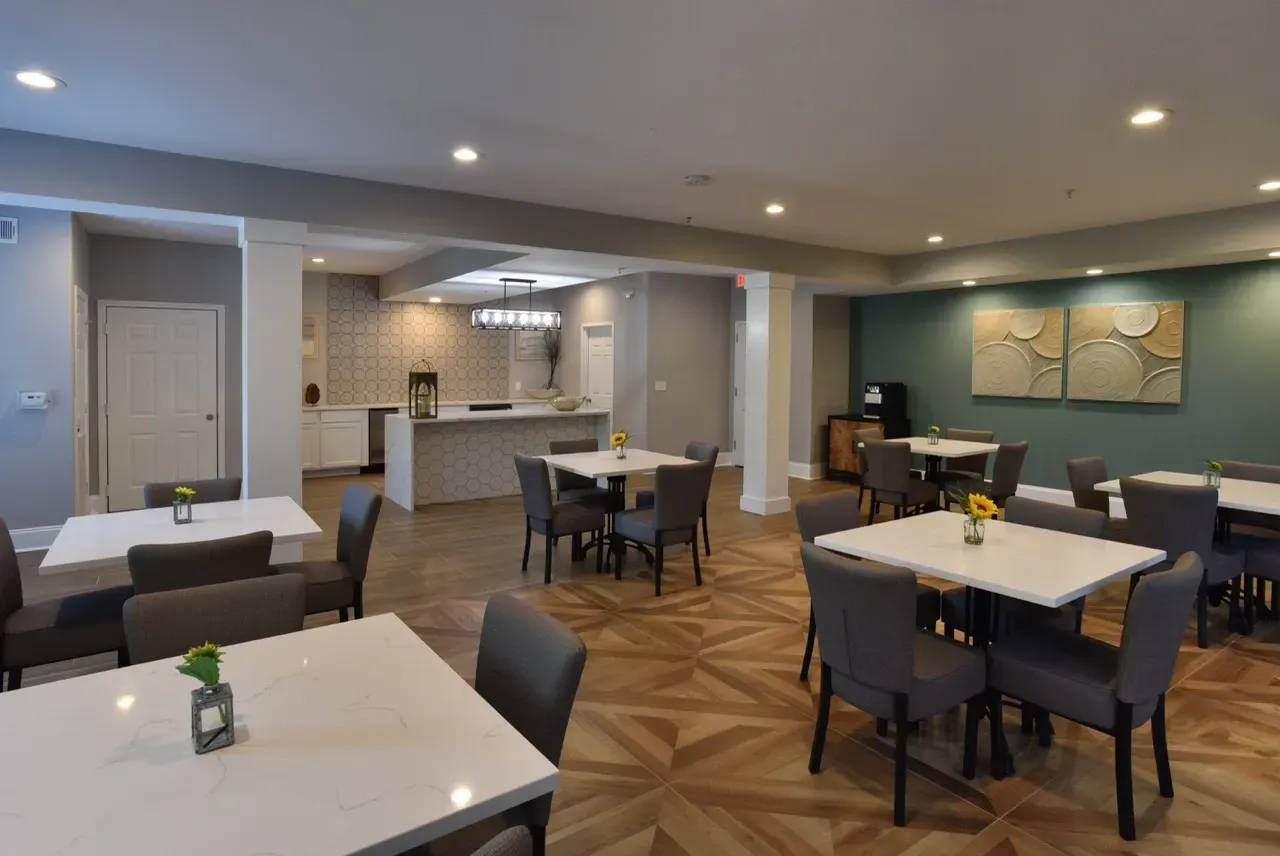 Indoor community lounge with white tables, gray chairs, and a kitchenette.
