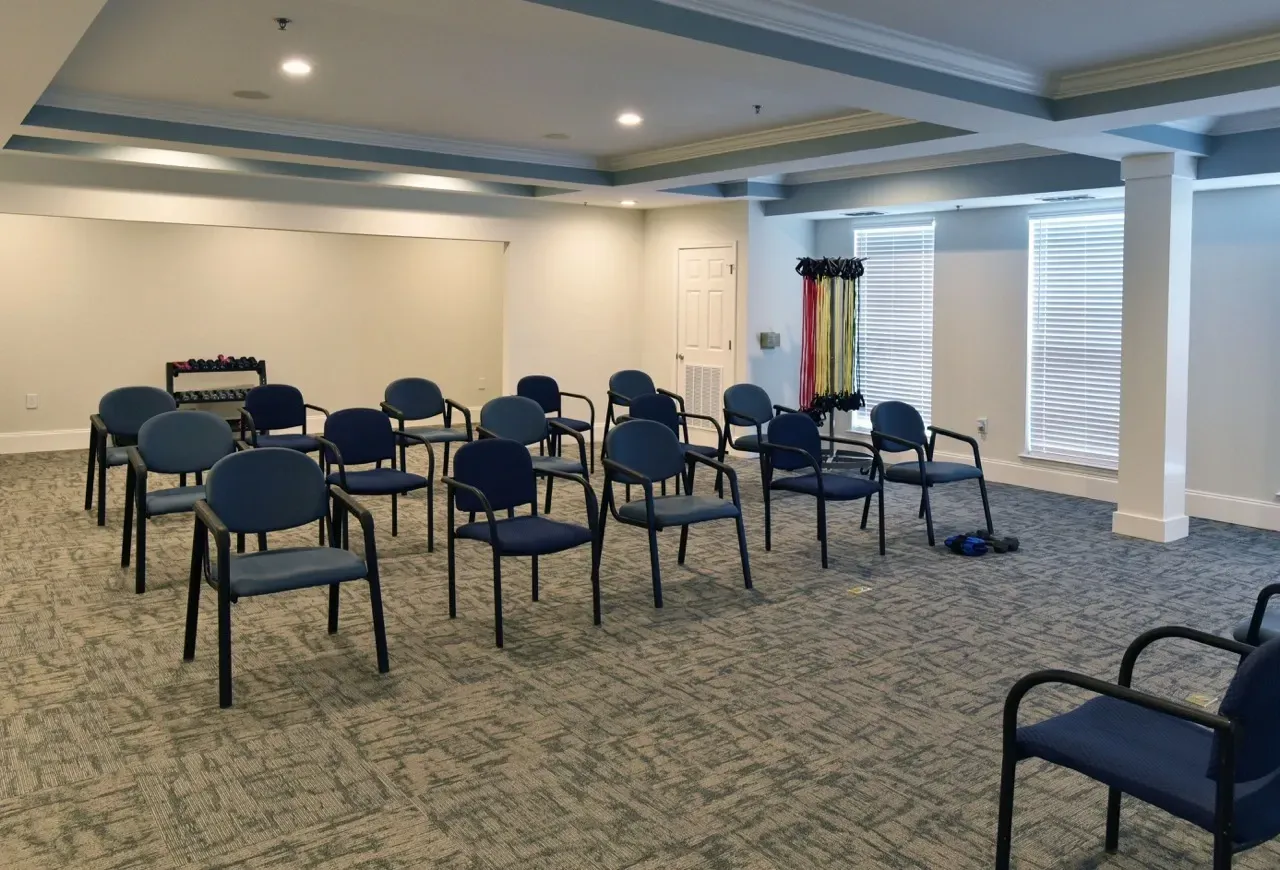 Indoor community room with blue chairs arranged in rows; dumbbells and resistance bands in the corner.
