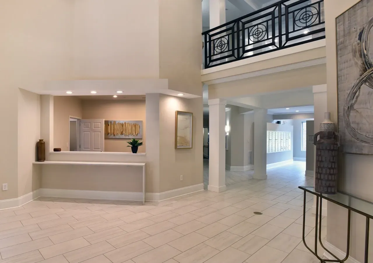 Open, well-lit apartment community lobby with a reception desk, columns, and upper-floor railing.