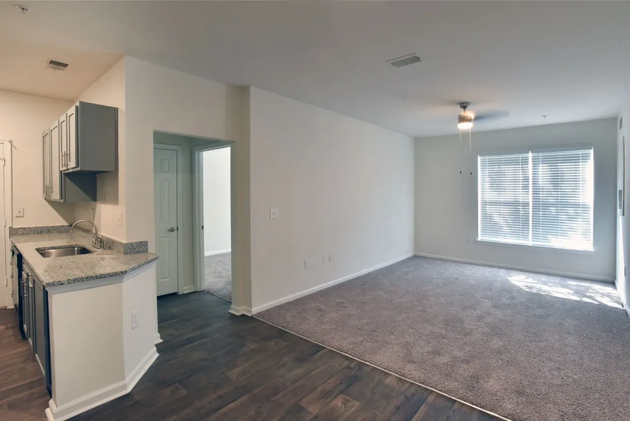 Open-concept kitchen with granite countertops and sink, adjacent to a carpeted living area with a large window.