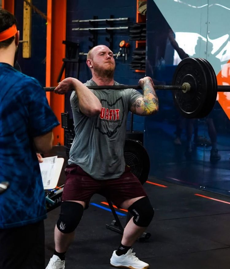 Weightlifter in UA Fit, a Columbus, OH gym, front-racking a barbell during a squat, with a coach watching nearby at the best gym in Columbus, OH.