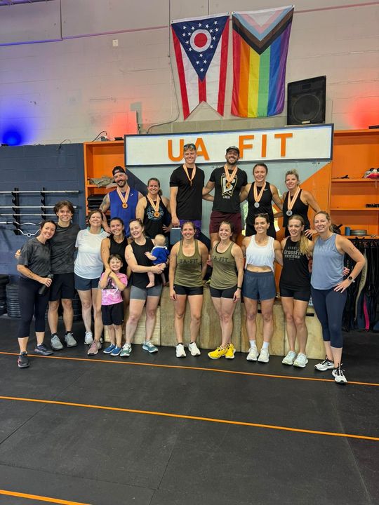 Group posing in UA Fit, a Columbus, OH gym, beneath UA FIT and rainbow flags, with blue seating in the background at the best gym in Columbus, OH.