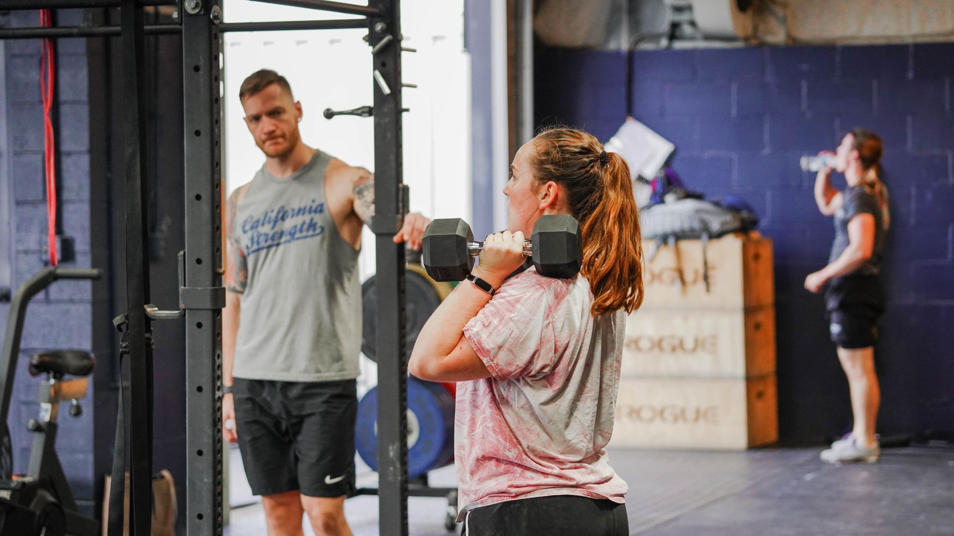 Gym class with a woman lifting dumbbells as a trainer watches and another person exercises in the background at the best gym in Columbus, OH.