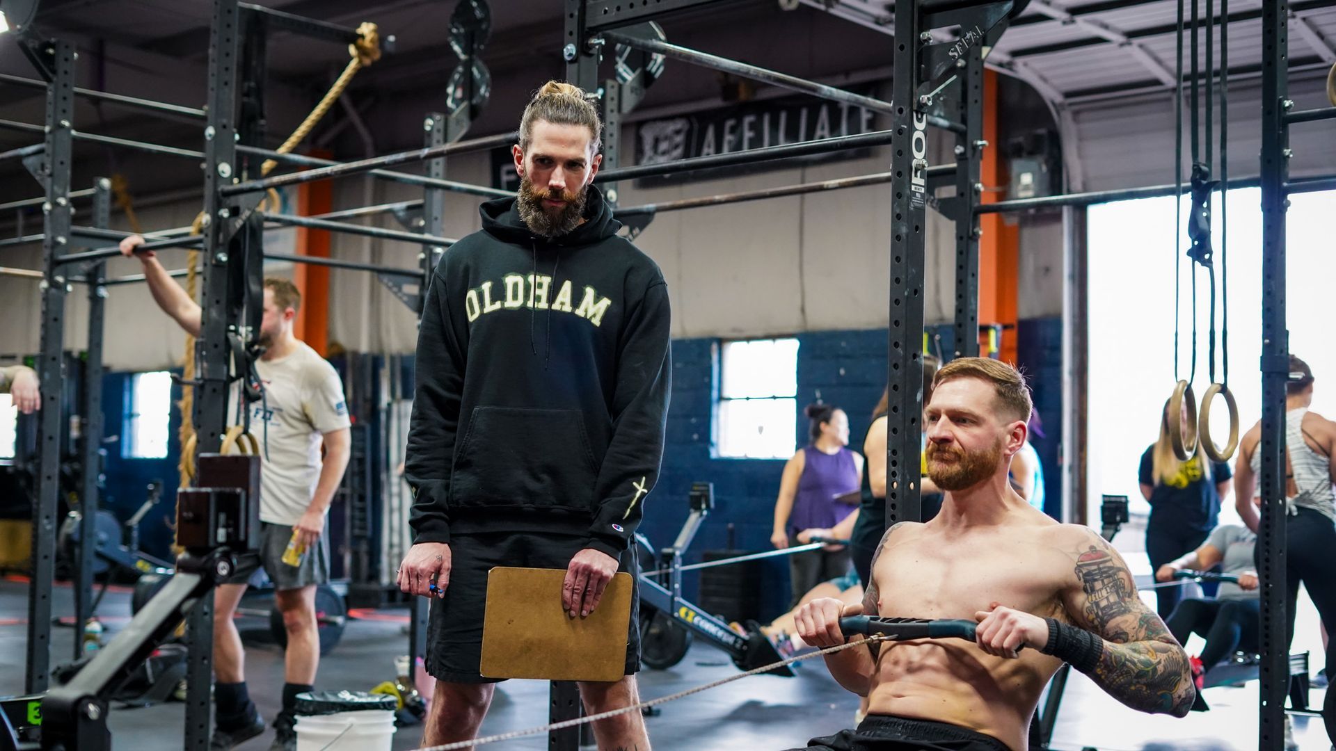 People exercising in a gym; one man sits on a rowing machine while others use rigs and pull-up bars at the best gym in Columbus, OH.