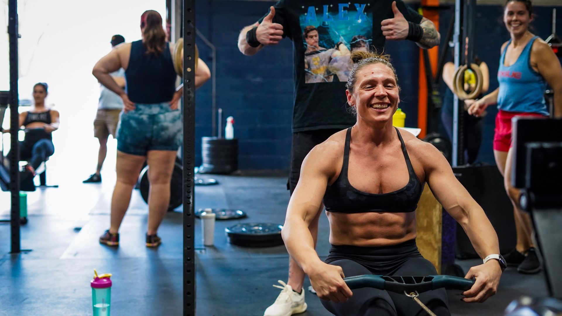Woman in a black sports bra smiling during a UA Fit, a Columbus, OH gym, workout with rowing machines and other exercisers behind her at the best gym in Columbus, OH.