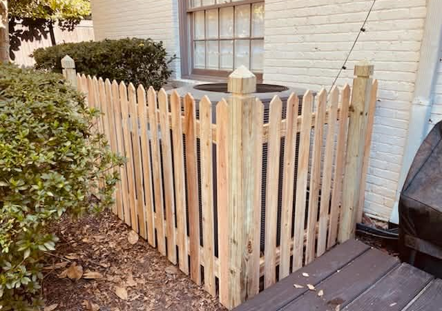 Wooden picket fence enclosing an AC unit next to a building and shrubbery.