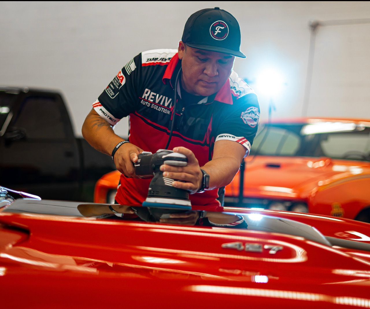 Man polishing a red car with a power buffer. Workshop setting with other vehicles.