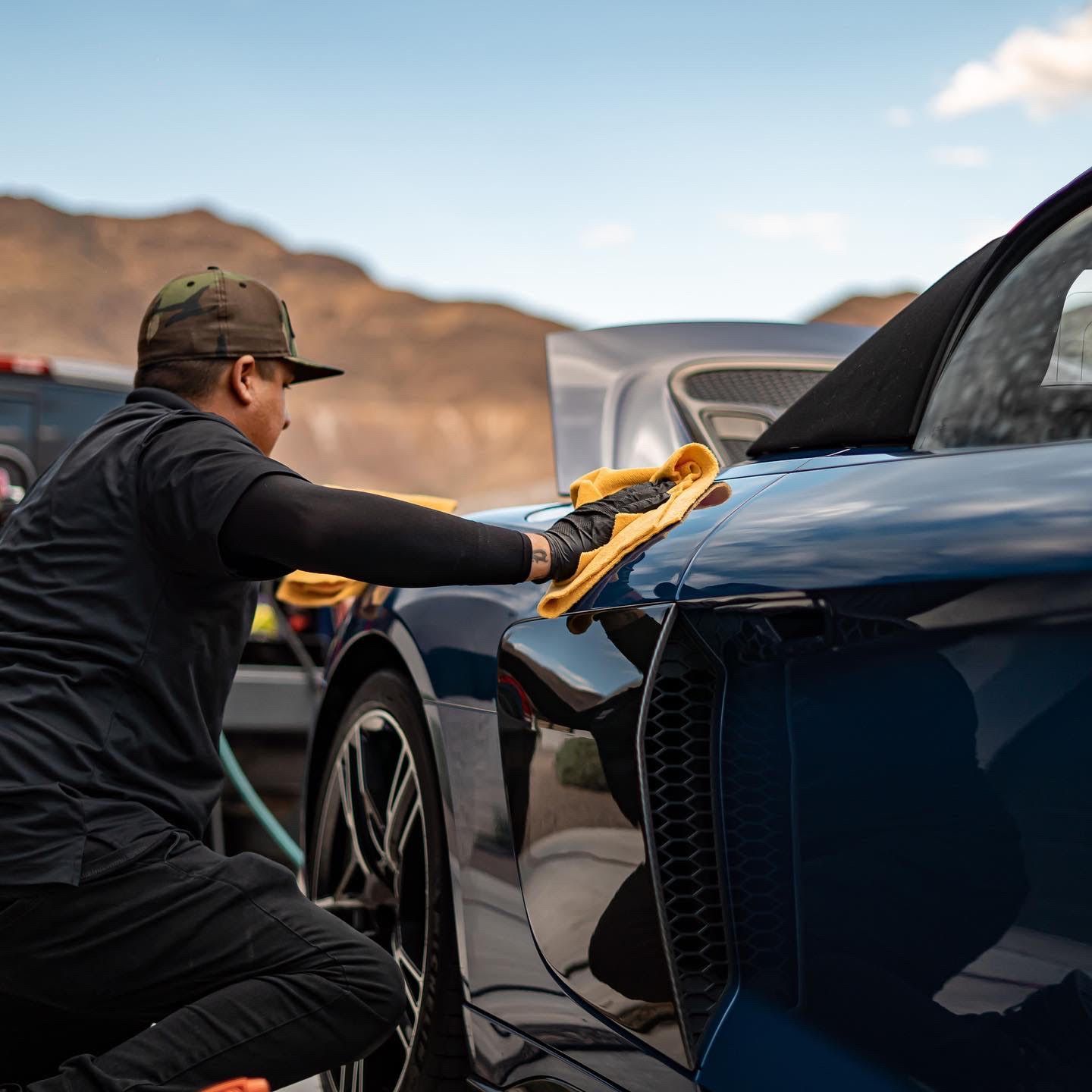 Man in black attire wiping down a blue car with a yellow cloth outdoors, mountains in the background.