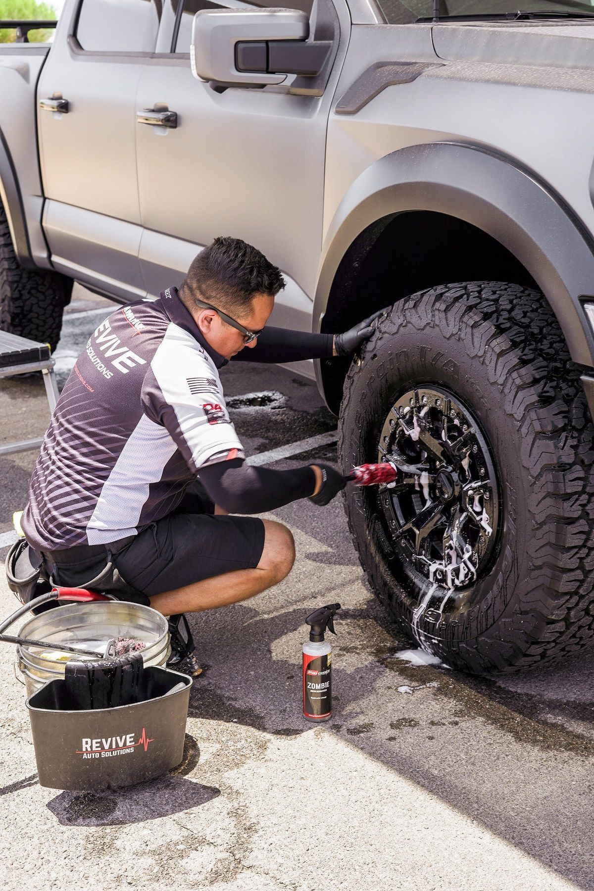 Person washing a truck tire with a brush and cleaning supplies outdoors.