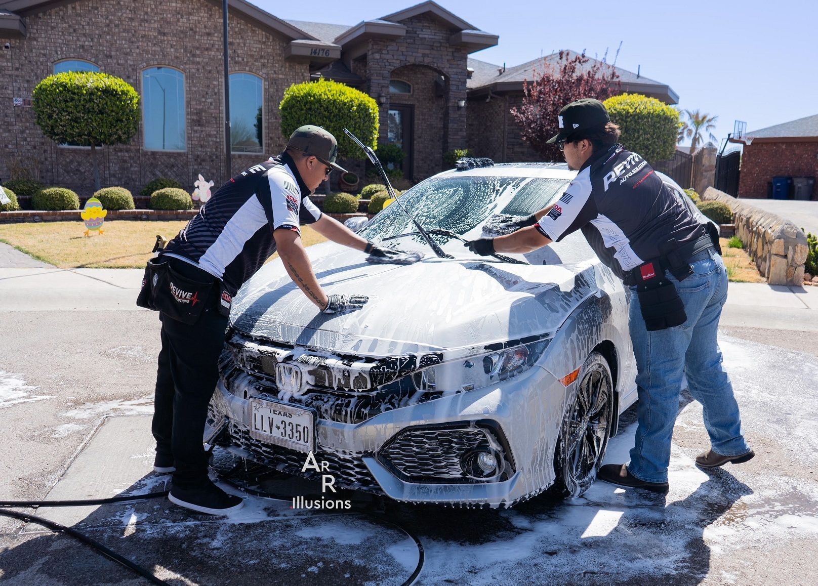 Two people washing a silver Honda Civic in front of a house, covered in soap suds.