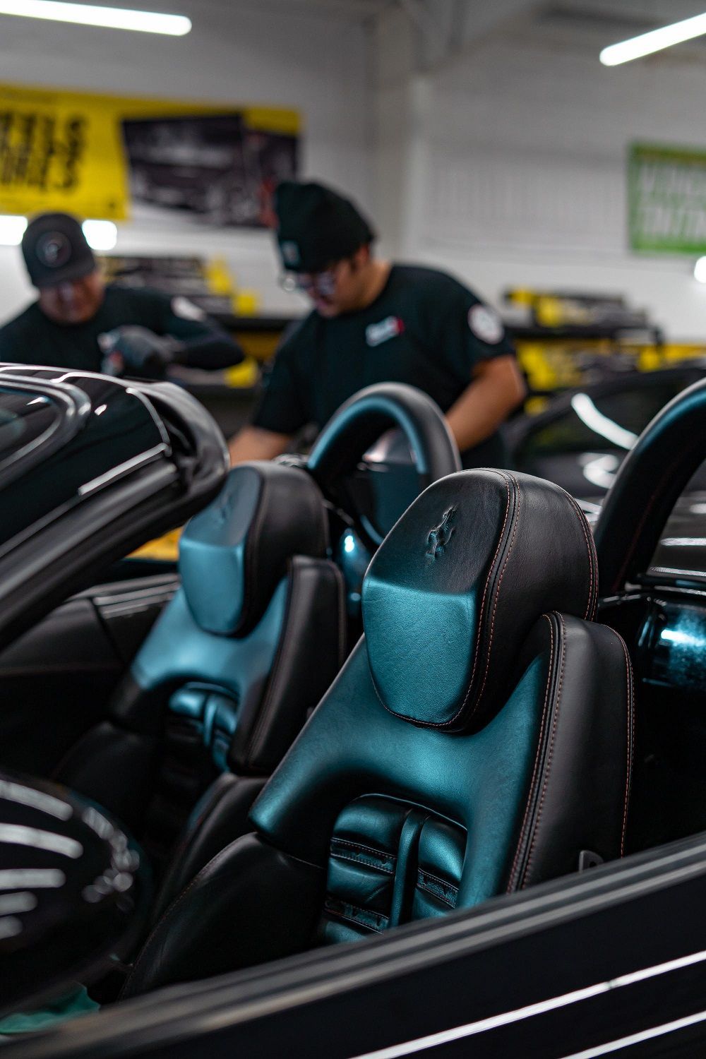 Black car interior with two people cleaning. Black leather seats with blue accents. Workshop setting.
