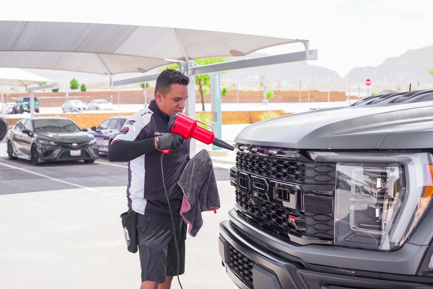 Man using a tool to clean the front of a gray pickup truck outdoors, with other cars and structures in the background.