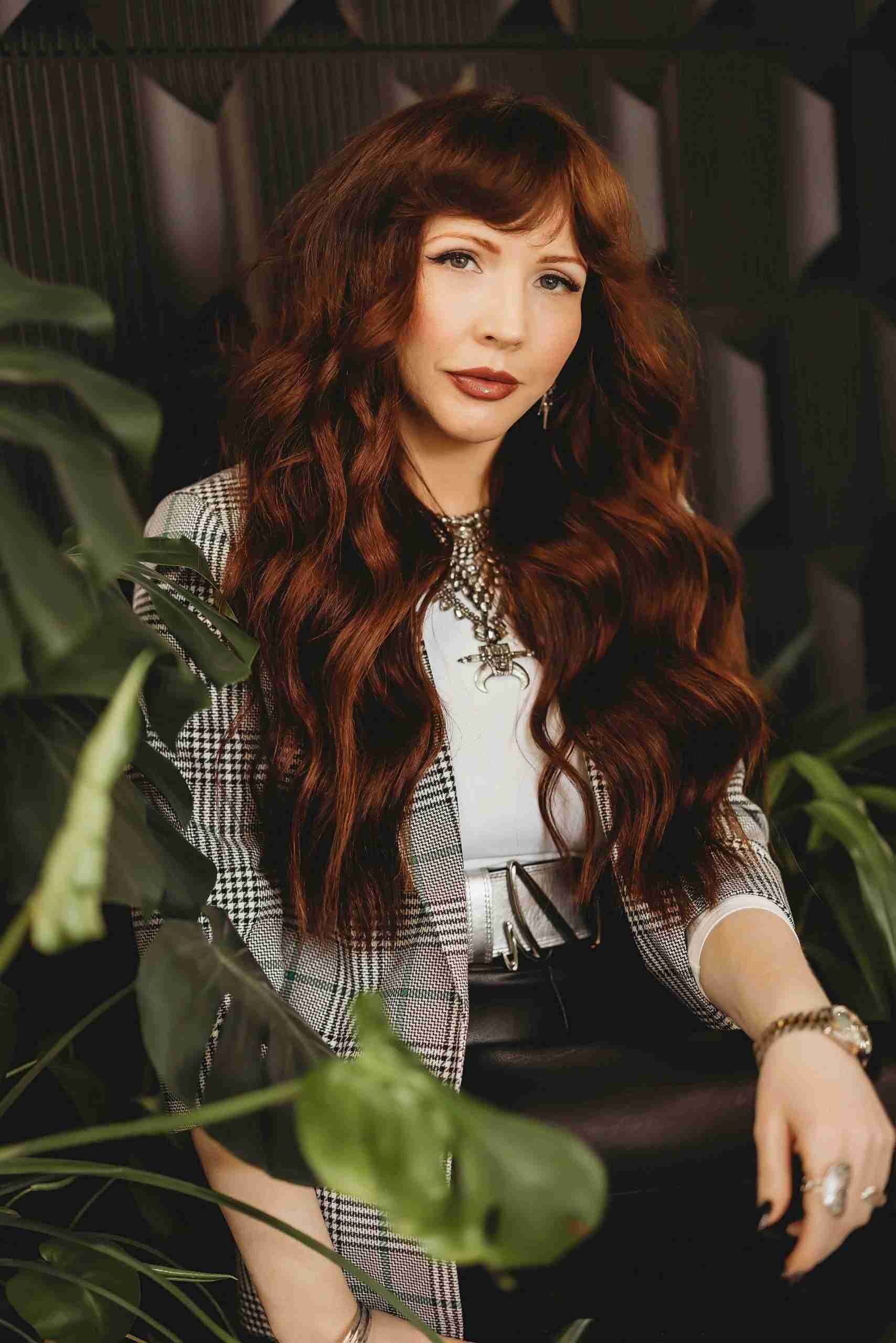 A woman with long red hair is sitting in a chair surrounded by plants.