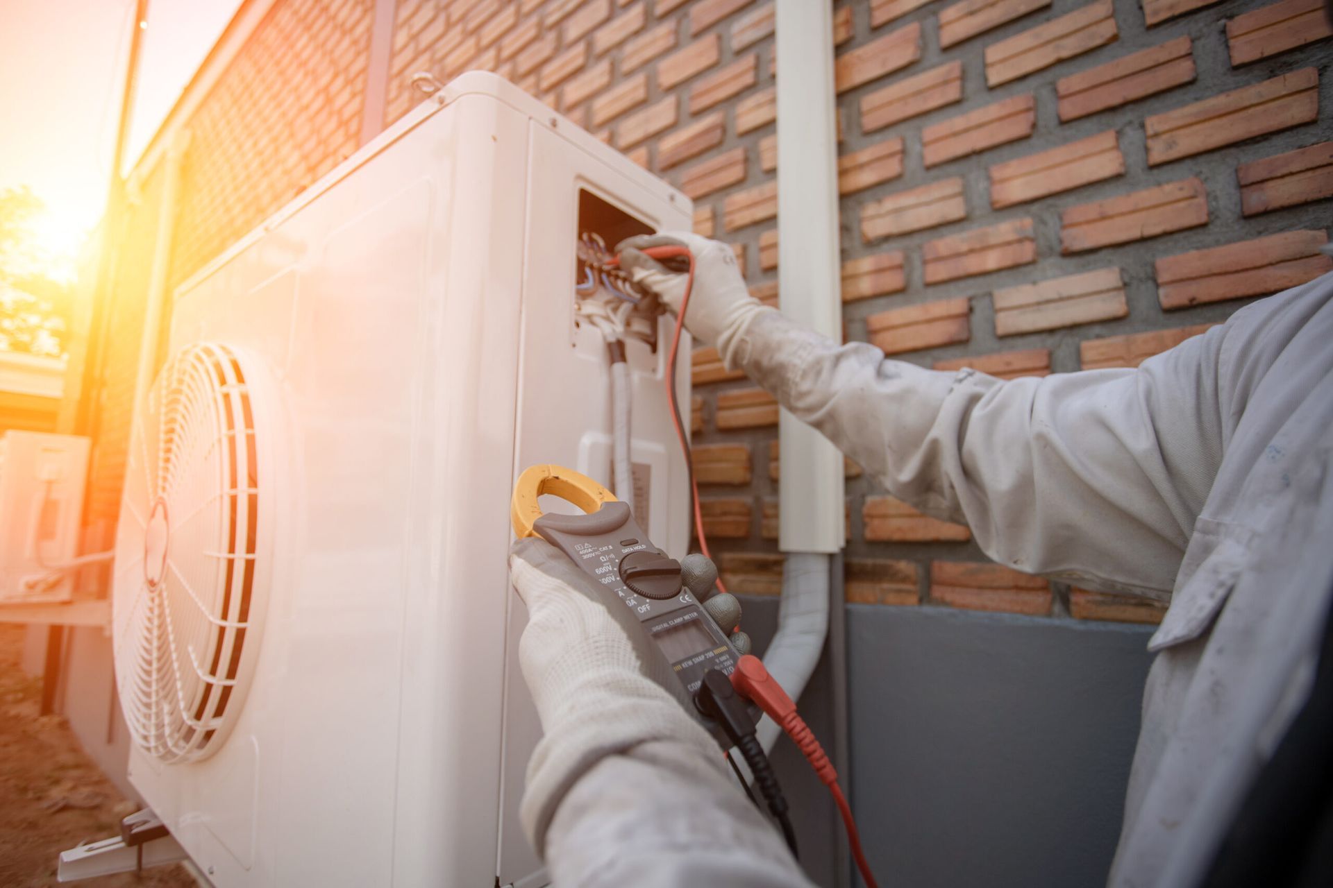 HVAC technician using a multimeter on an outdoor air conditioning unit next to a brick wall.