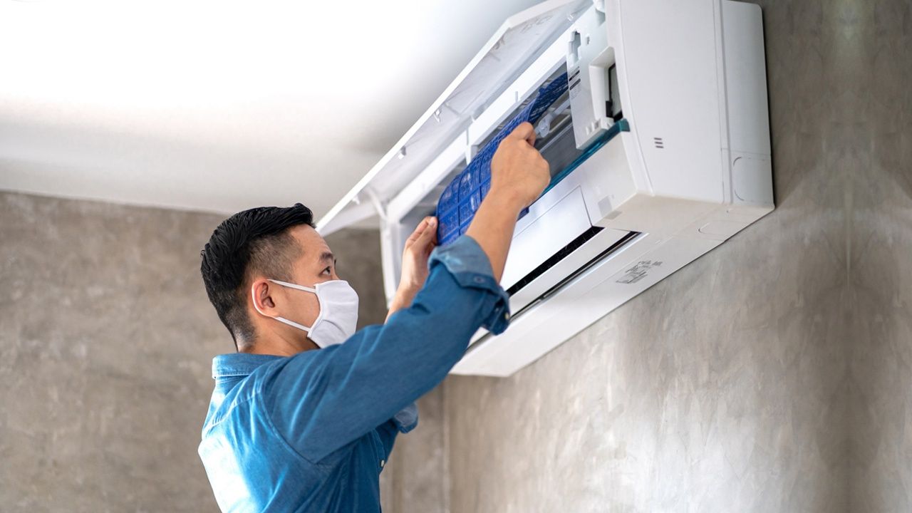 A technician wearing a face mask services a wall-mounted air conditioning unit.
