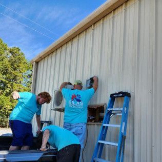 Three people install an AC unit on a corrugated metal building. Blue shirts, ladder, sunny day.