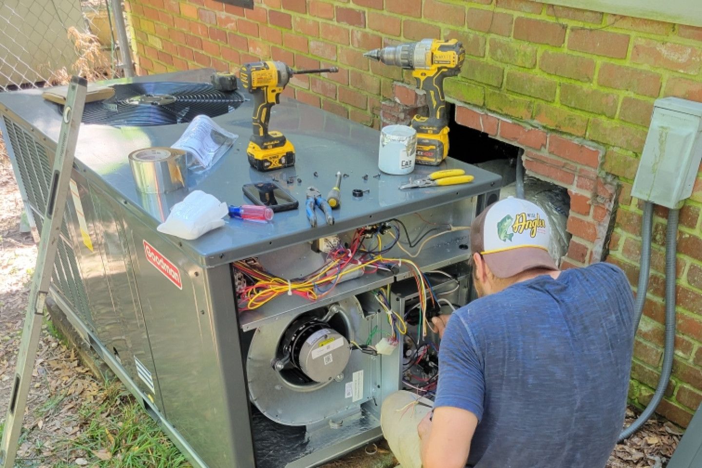 HVAC technician repairs an outdoor furnace. Tools and parts are scattered on top. Brick wall is adjacent.